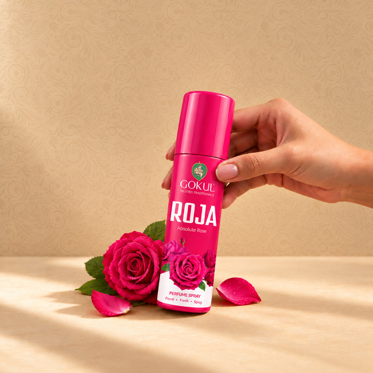 A hand holds a bottle of Gokul Roja Rose perfume spray against a beige patterned background, with red roses and petals on a beige surface below.