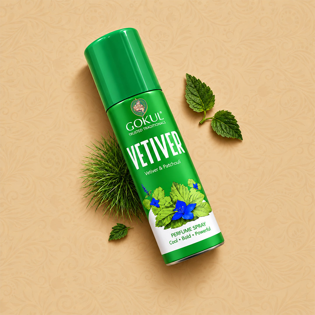 A green aerosol can of Gokul Vetiver Perfume Spray, styled diagonally on a textured beige background with decorative vetiver grass, mint leaves, and a blue flower.